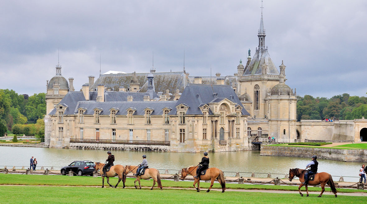 A beautiful French castle next to water with people riding horses in the foreground